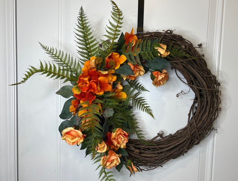 May include: A brown grapevine wreath decorated with orange and yellow flowers and green ferns.