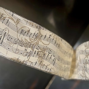May include: Cream-colored ribbon with musical notes and decorative floral accents. The ribbon has a silver glitter edge and is curled, showing the pattern of the musical score. The ribbon is likely used for crafts or gift wrapping.