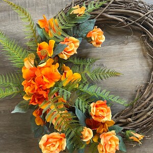 May include: A brown grapevine wreath decorated with orange and yellow silk flowers and green fern leaves.