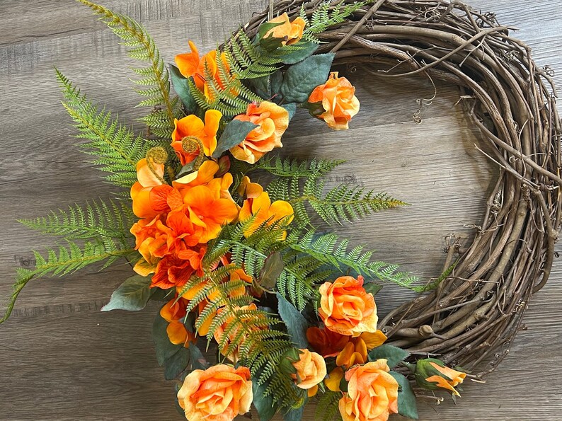 May include: A brown grapevine wreath decorated with orange and yellow silk flowers and green fern leaves.