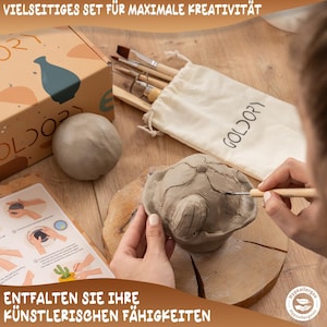 May include: A set of pottery tools and clay for crafting. The tools include a wooden handle with a metal tip, a small wooden tool, and a small wooden tool with a metal tip. The clay is in a bag with the word "GOLDORY" printed on it. The text "VIELSEITIGES SET F&Uuml;R MAXIMALE KREATIVIT&Auml;T" is printed on a box in the background. The text "ENTFALTEN SIE IHRE K&Uuml;NSTLERISCHEN F&Auml;HIGKEITEN" is printed on a white surface in the foreground.