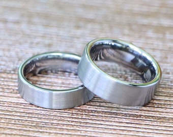 May include: Two silver wedding bands, one on top of the other, on a wooden surface.
