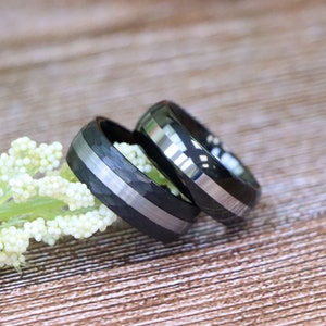 May include: Two black and silver wedding bands. One band has a brushed finish and the other has a carbon fiber pattern.