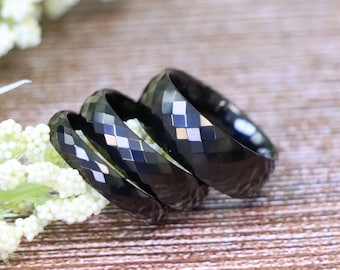 May include: Three black faceted rings of varying sizes are displayed. The rings have a geometric pattern and are set against a wooden surface with white flowers in the background. The rings are likely wedding or fashion rings.
