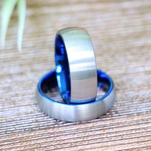 May include: Two silver-colored rings with a brushed metal finish. The interior of each ring is a vibrant blue. The rings are displayed on a wooden surface, showcasing their contrasting colors and textures. The rings are likely wedding bands or fashion accessories.