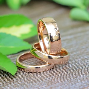 May include: Three gold wedding bands are stacked together, reflecting the surrounding environment. The rings have a polished finish and a rounded shape. The background includes green leaves and a wooden surface.