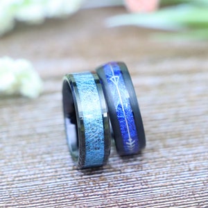 May include: Two black wedding bands. One ring features a light blue, textured inlay. The other ring has a dark blue inlay with a silver arrow design. Both rings are set against a blurred wooden background.