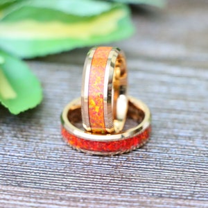 May include: Two gold-colored rings with a vibrant orange and yellow opal inlay. The rings have a polished finish and a grooved design on the edges. The opal has a speckled appearance, creating a unique look.
