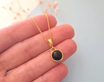 Op de afbeelding: Een goudkleurige ketting met een fijne schakelketting en een ronde hanger met een gladde, gepolijste zwarte steen in een goudkleurige metalen zetting. De hanger hangt aan een eenvoudig oogje, wat zorgt voor een strak en klassiek sieraad.