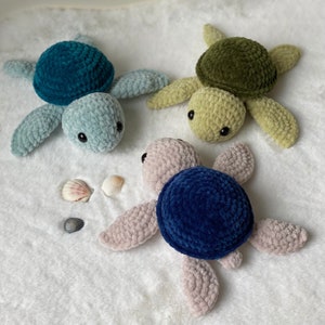Könnte beinhalten: Drei Plüschschildkröten in Blau-, Grün- und Rosatönen mit schwarzen Augen. Die Schildkröten sind auf einer weißen, flauschigen Oberfläche angeordnet, mit drei Muscheln in der Nähe. Die Schildkröten sind handgefertigt.