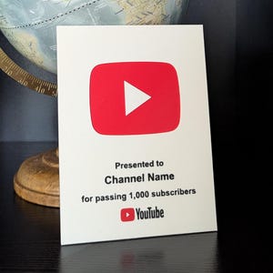 May include: A classic globe with a gold-coloured frame and base, alongside a white YouTube play button award. The award displays a red play button with a white triangle and the text: "Presented to Channel Name for passing 1,000 subscribers."