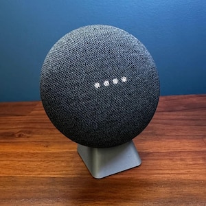 May include: A dark gray smart speaker with a fabric cover, resting on a gray stand. Four white lights are visible on the front. The speaker sits on a wooden surface against a blue wall.