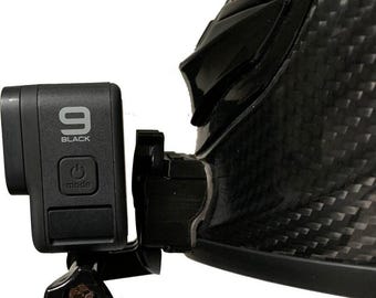 May include: Black GoPro 9 camera attached to a black helmet. The camera is mounted with a black bracket and features the word "BLACK" and the number "9". The camera has a mode button and a power button.