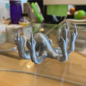 Könnte beinhalten: Eine silberne Oktopus-Figur mit mehreren Armen, die jeweils in einer Hand enden, die den Mittelfinger zeigt. Der Oktopus befindet sich auf einer reflektierenden Oberfläche, mit anderen Objekten im Hintergrund.