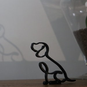 May include: A black metal wire sculpture of a dog sitting with its head tilted to the side. The sculpture is on a wooden surface.