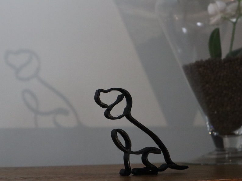 May include: A black metal wire sculpture of a dog sitting with its head tilted to the side. The sculpture is on a wooden surface.