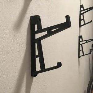 Puede incluir: Tres soportes para bicicletas de metal negro montados en una pared blanca. Los soportes están diseñados para sujetar una bicicleta por la rueda delantera.