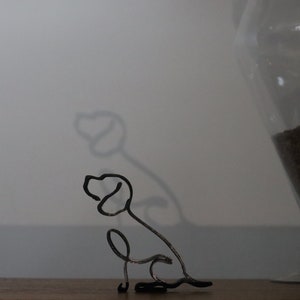 May include: A black metal wire sculpture of a dog sitting with its head turned to the side. The sculpture is on a wooden surface.