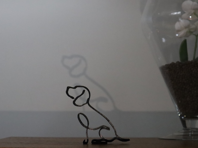May include: A black metal wire sculpture of a dog sitting with its head turned to the side. The sculpture is on a wooden surface.