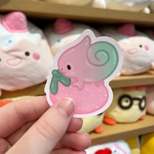 Può includere: Un adesivo rosa e verde con un camaleonte dei cartoni animati che tiene una fragola. Il camaleonte ha un corpo rosa, occhi verdi e una coda verde. La fragola è rosa con foglie verdi.
