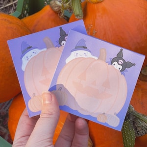 May include: Two purple and white Halloween themed sticky notes with a pumpkin and a ghost wearing a witch hat. The ghost is holding a black cat with a witch hat. The notes are in front of a pile of pumpkins.