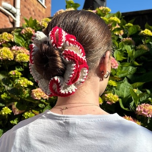 May include: A white and red crocheted hair scrunchie with a ruffled design. The scrunchie is worn in a woman's hair.