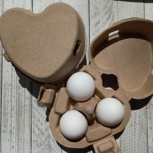 May include: A heart-shaped, brown cardboard egg carton holding three white eggs. The carton is open, revealing the eggs nestled in their individual compartments. The carton is on a light-colored, wooden surface.