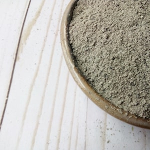 May include: A brown ceramic bowl filled with a grey, granular substance. The bowl is in the upper right corner against a light-coloured, wooden surface with visible grain. The substance appears to be a fine powder.