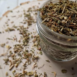 May include: A clear glass jar filled with dried herbs, with some spilled onto a light-colored wooden surface. The herbs are a mix of green and brown, suggesting a natural product. The jar is in focus, with the background slightly blurred.