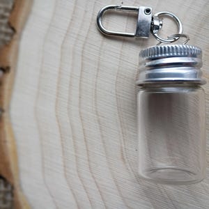 May include: A small, clear glass vial with a silver-colored metal screw-on cap and a silver-colored metal clasp. The vial is empty and sits on a light-colored wooden surface.