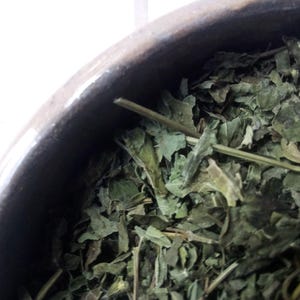 May include: Close-up of dried green leaves filling a dark brown ceramic bowl. The leaves have a textured appearance and vary in size and shape. The bowl's rim is visible, with a slightly glossy finish.