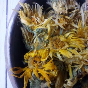 Könnte beinhalten: Nahaufnahme von getrockneten gelben Ringelblumen in einer dunklen Keramikschale. Die Blüten haben eine leuchtend gelbe Farbe mit einigen Grün- und Brauntönen. Die Blütenblätter sind zart und strukturiert, mit einem natürlichen, organischen Aussehen.