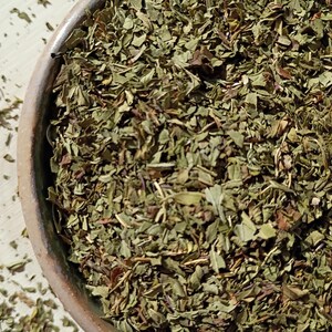 May include: Close-up of dried green herbs in a brown ceramic bowl. The herbs are finely chopped and have a mix of green and brown hues. Some herbs are scattered outside the bowl on a light-colored surface. The image suggests culinary or herbal use.