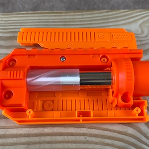 May include: A bright orange plastic toy blaster component featuring a rectangular housing with ribbed detailing, a central cylindrical chamber containing a metallic silver rod, and a circular barrel opening on the right side with visible screws.