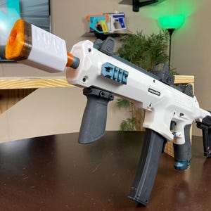 May include: A white and black toy gun with a black magazine and a teal colored grip. The gun has a white barrel extension with the word "WORKER" printed on it.
