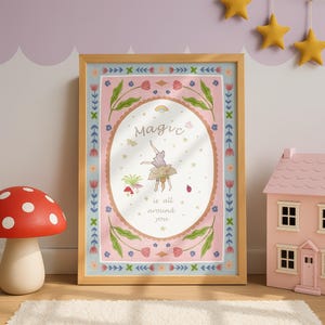 May include: A framed rectangular wall art print featuring a pink border with floral motifs and a central oval containing a person doing a handstand. The text reads Magic is all around you, surrounded by small illustrations of a rainbow, stars, and a ladybug.