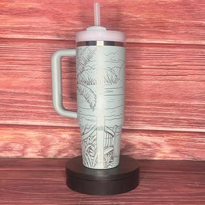 K&ouml;nnte beinhalten: Ein hellblauer isolierter Becher mit Griff, Deckel und Strohhalm. Der Becher zeigt eine Strandszene mit Palmen, einem Zaun und der Sonne. Der Griff und der Deckel sind hellgrau. Der Becher ist etwa 25 cm hoch.