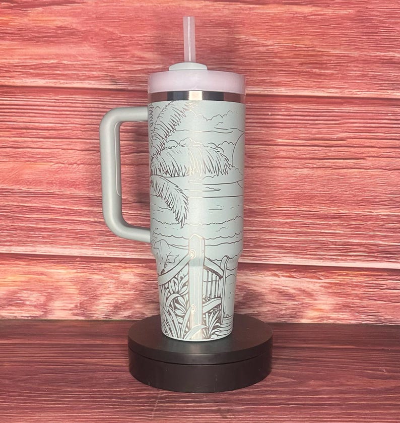 K&ouml;nnte beinhalten: Ein hellblauer isolierter Becher mit Griff, Deckel und Strohhalm. Der Becher zeigt eine Strandszene mit Palmen, einem Zaun und der Sonne. Der Griff und der Deckel sind hellgrau. Der Becher ist etwa 25 cm hoch.