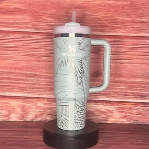 K&ouml;nnte beinhalten: Ein hellblauer Stanley-Becher mit Griff und rosa Deckel. Der Becher zeigt eine Strandszene mit Palmen und einem Zaun. Das Stanley-Logo ist sichtbar. Enth&auml;lt einen wiederverwendbaren Strohhalm.