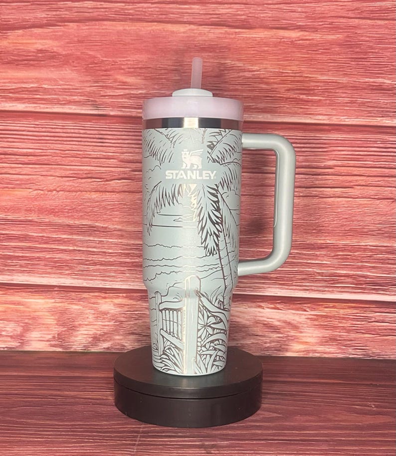 K&ouml;nnte beinhalten: Ein hellblauer Stanley-Becher mit Griff und rosa Deckel. Der Becher zeigt eine Strandszene mit Palmen und einem Zaun. Das Stanley-Logo ist sichtbar. Enth&auml;lt einen wiederverwendbaren Strohhalm.