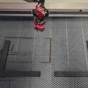 May include: A laser cutter in operation, with a red cutting head and two black L-shaped pieces. The cutting bed has a honeycomb pattern. The machine is cutting a square shape.
