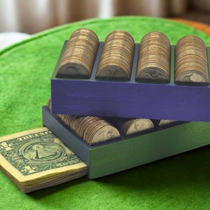 May include: Two wooden coin holders, one purple and one green, stacked on top of each other. Each holder contains rolls of coins. A stack of dollar bills is partially visible beneath the green holder, with the text "ONE DOLLAR" visible.
