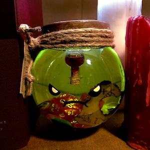May include: A glass jar with a green liquid inside. The jar has a cork stopper and a small glass vial inside. The jar is decorated with a pirate map and a green, red, and yellow dice. The jar has a label that says "NOT FOR CONSUMPTION".