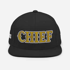 Puede incluir: Gorra de béisbol negra con un diseño bordado en oro con la palabra "CHIEF" en la parte delantera.