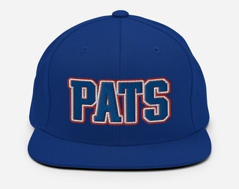 May include: A royal blue baseball cap featuring the word PATS embroidered in large, bold letters across the front. The lettering is filled with blue thread and outlined in white and red, creating a distinct layered effect on the structured crown.