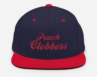 Könnte beinhalten: Eine marineblaue Baseballkappe mit flachem rotem Schirm und einem roten Knopf oben. Auf der Vorderseite ist der Schriftzug Peach Clobbers in roter Schreibschrift eingestickt. Die Kappe hat rote Ösen an den Seitenteilen zur Belüftung.