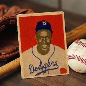 May include: Vintage baseball card featuring a black and white photo of a baseball player wearing a blue cap with a white "B" on it. The player is smiling and wearing a white baseball uniform with the word "Dodgers" written in blue cursive. The card has a red background.