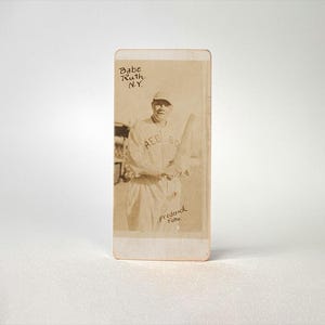 May include: A vintage baseball card featuring Babe Ruth, a baseball player for the New York Yankees. The card is black and white and shows Ruth holding a baseball bat. The text on the card reads "Babe Ruth, N.Y." and "Frederick  Pabe".