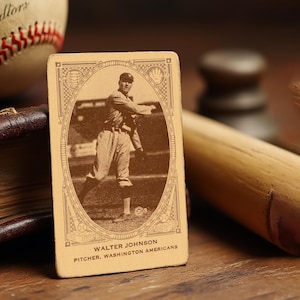 May include: Vintage baseball card featuring Walter Johnson, pitcher for the Washington Americans. The card is in sepia tones, with an oval portrait of Johnson in action. A baseball and bat are also visible.