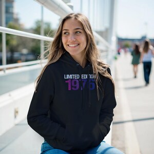 Könnte beinhalten: Schwarzer Kapuzenpullover mit dem Text "LIMITED EDITION 1976" in Weiß, Lila und Blau. Der Hoodie hat eine Kapuze mit Kordelzug und eine Vordertasche. Die Person trägt eine blaue Jeans.